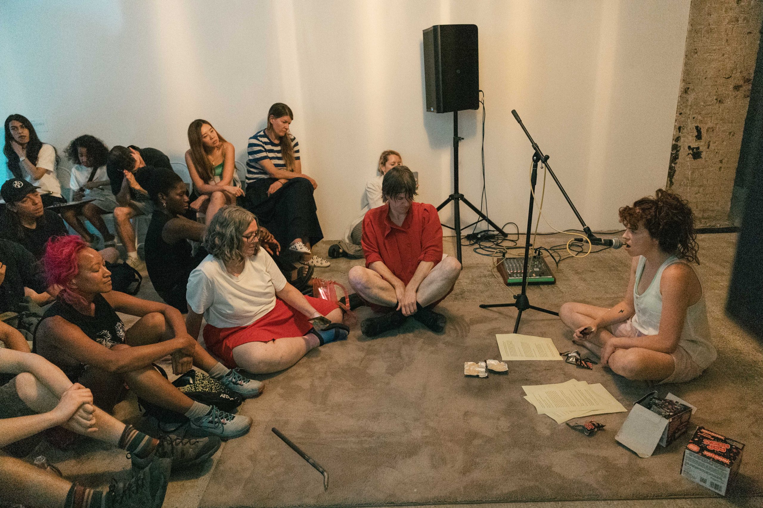 A woman sits on a grey rug talking into a microphone with pages of white paper in front of her. A crowd sits on the floor in front of her.