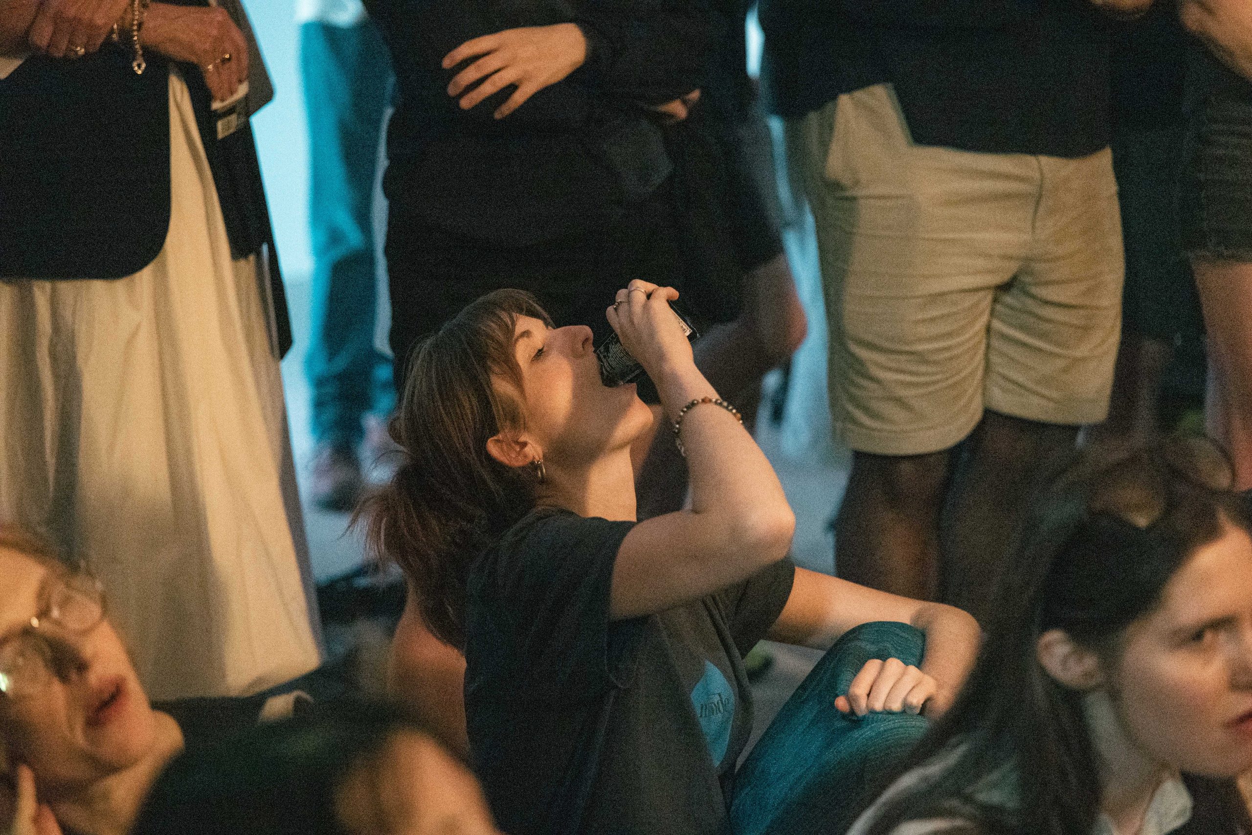 A woman sits in the middle of a crowd pouring something into her mouth.