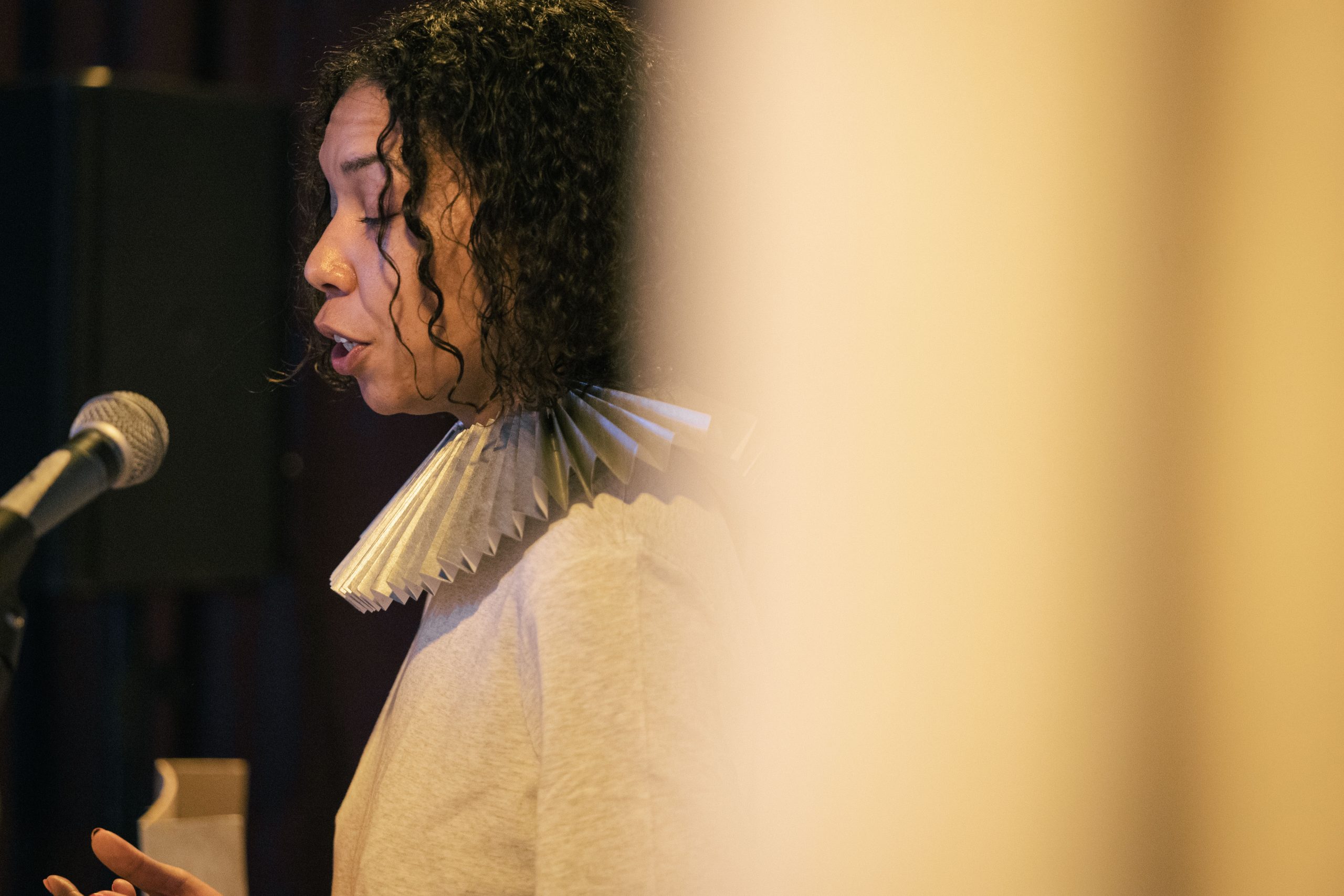 A close-up of a woman talking into a microphone behind a white curtain.