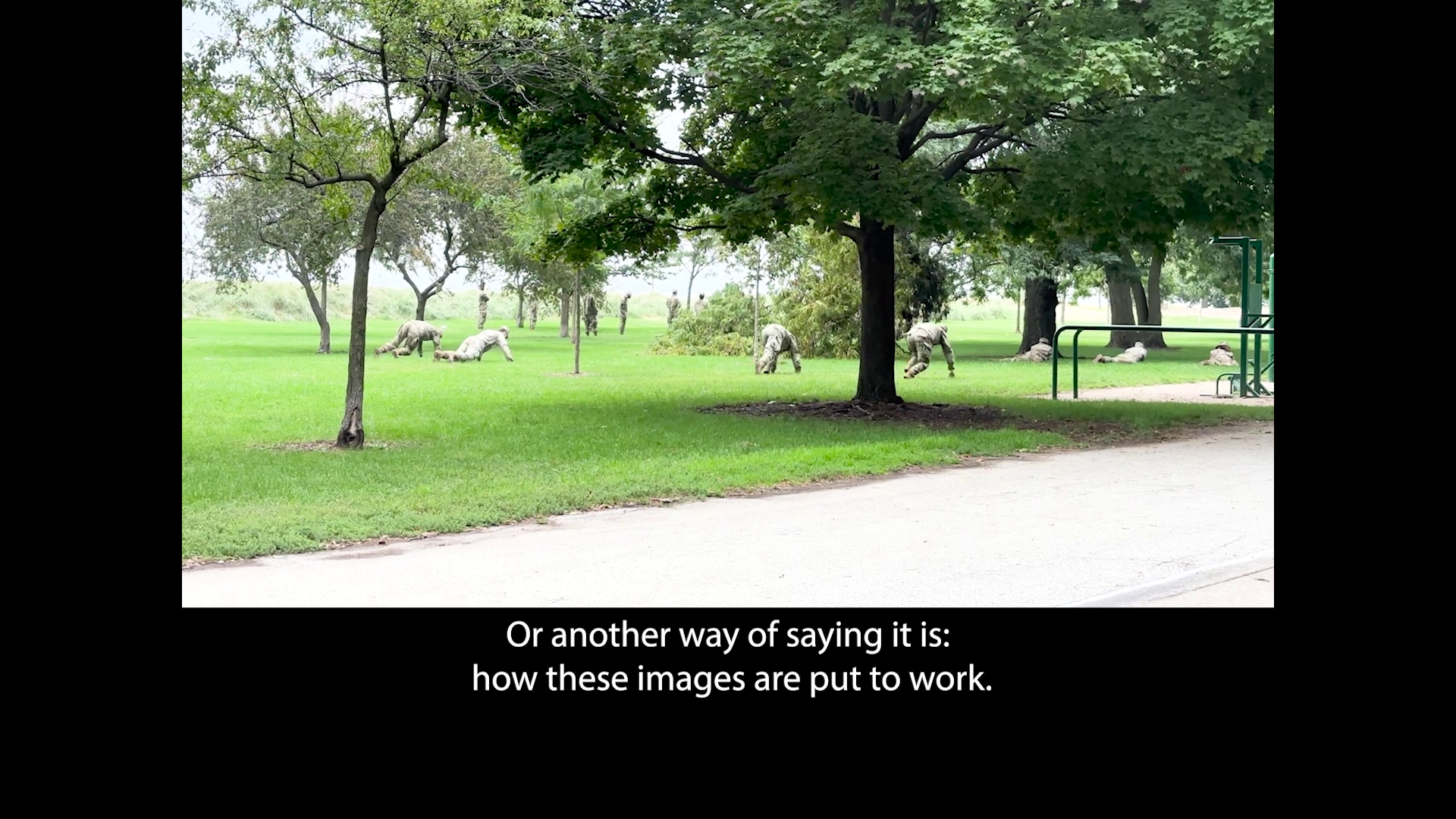 Soldiers lower to the ground, doing a training exercise in a green space in a park, framed by a u-shaped black border that makes the image look almost as if it’s on a stage.Beneath the image, a caption appears in the black space, “Or another way of saying it is, how these images are put to work.”