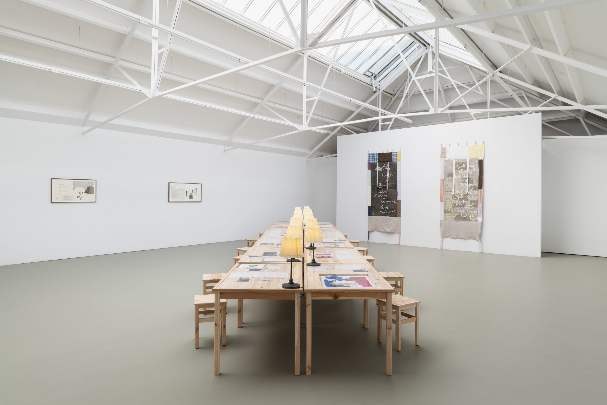 Art gallery interior featuring textile artworks on walls and a long table with chairs, illuminated by table lamps.
