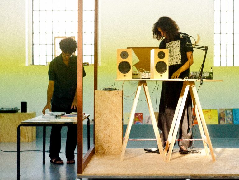 A man stands behind a table of books next to raised platform holding a woman standing behind a wooden trestle table holding two speakers.