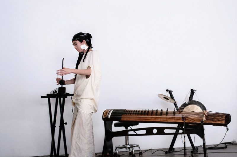 Woman stands in front of a white wall playing a theremin. A chinese instrument is behind her.