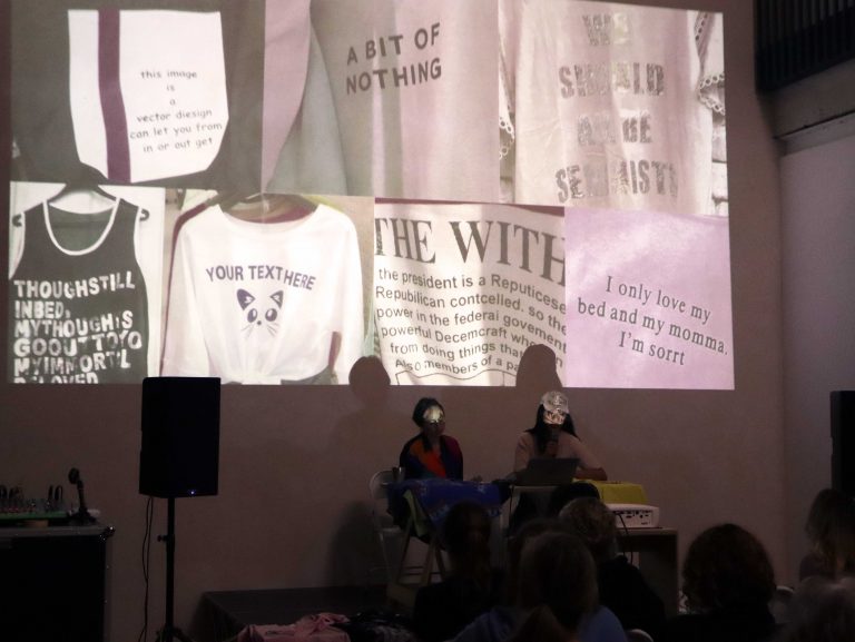 A large projection of different t shirts in colours of pink, black and white with different writing on them. Two people sit at a desk in front of the projection and a large black speaker stands on the left.